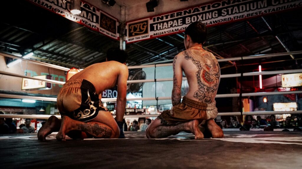 Two tattooed Muay Thai fighters kneeling in a boxing ring at Thapae Boxing Stadium.